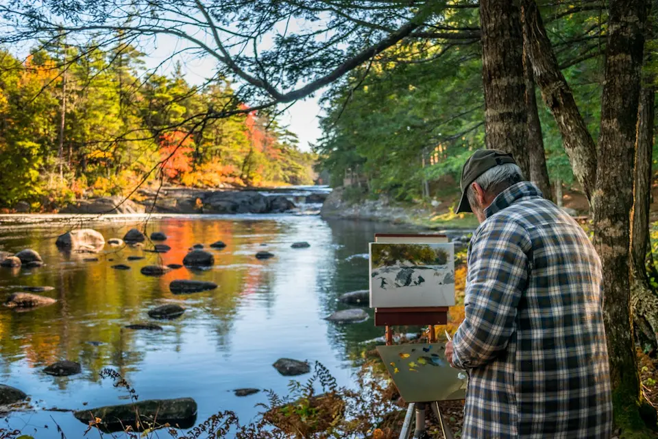 Unrecognizable painter with easel against river and autumn trees